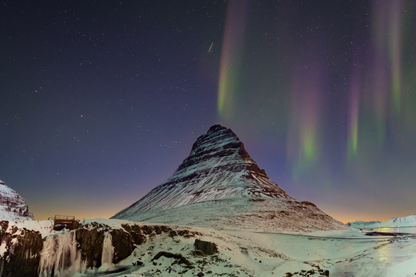 Pale green and purple northern lights rays dance over a cone-shaped mountain in the snowy landscape. In foreground, a small frozen waterfall