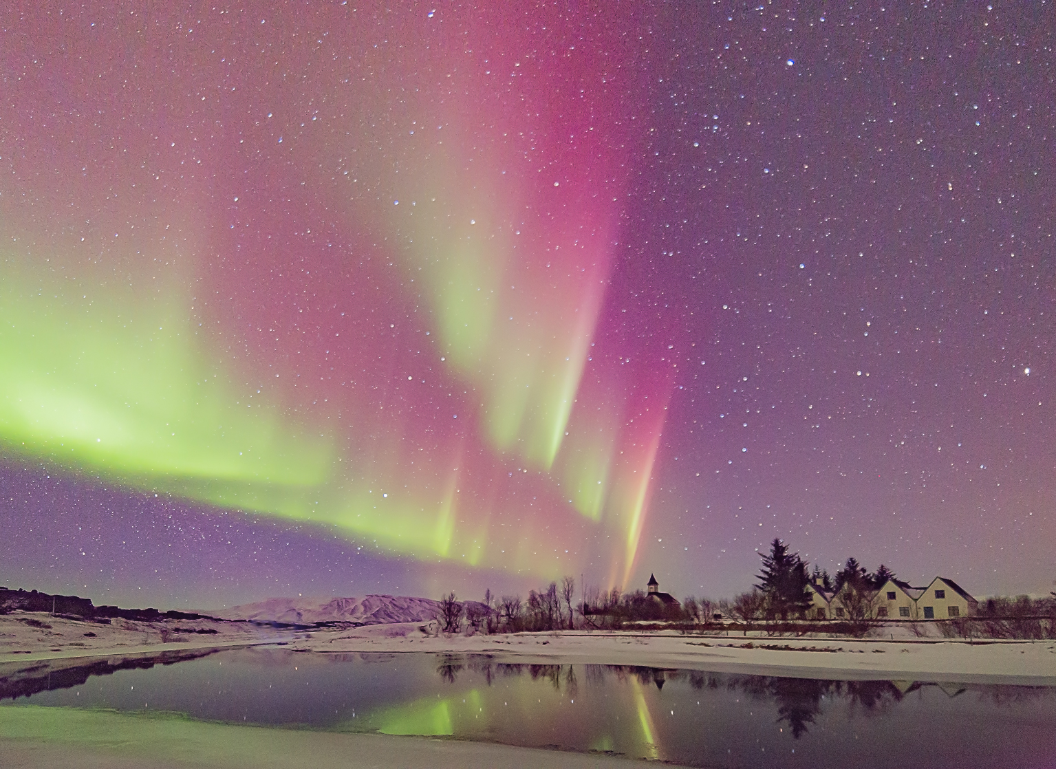 bright pink and green northern lights reflect in a pond, a church can be seen in backdrop in the snowy landscape