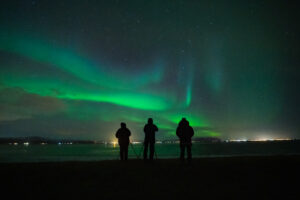 Three people with cameras and tripods watch the northern lights dance in the night sky