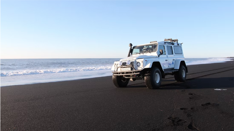 White superjeep 6 seater vehicle driving on a black sand beach oceanside