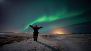 A woman has her picture taken in front of a beautiful northern lights display on a dark winter night. Plenty of snow on the ground