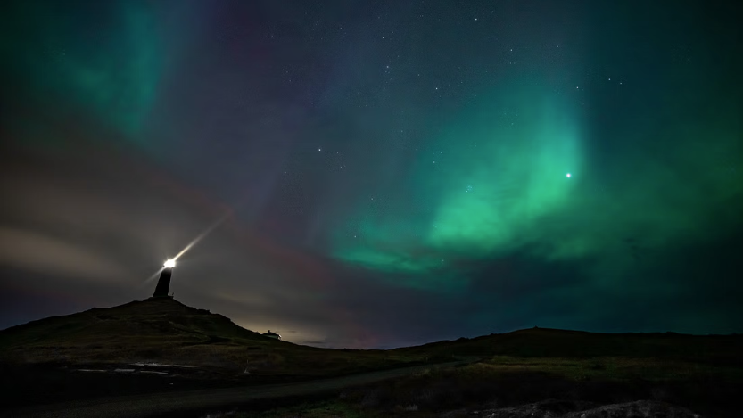 Green northern Lights and stars over a hill, a lighthouse is visible in backdrop