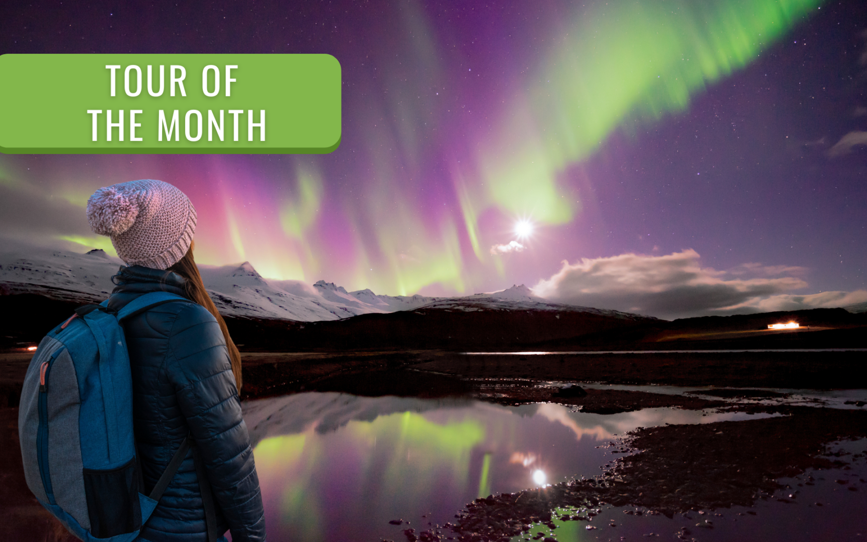 A woman with a backpack and woolly hat gazing at a spectacular purple and green aurora borealis reflected in a still lake, with snow-capped Icelandic mountains in the background