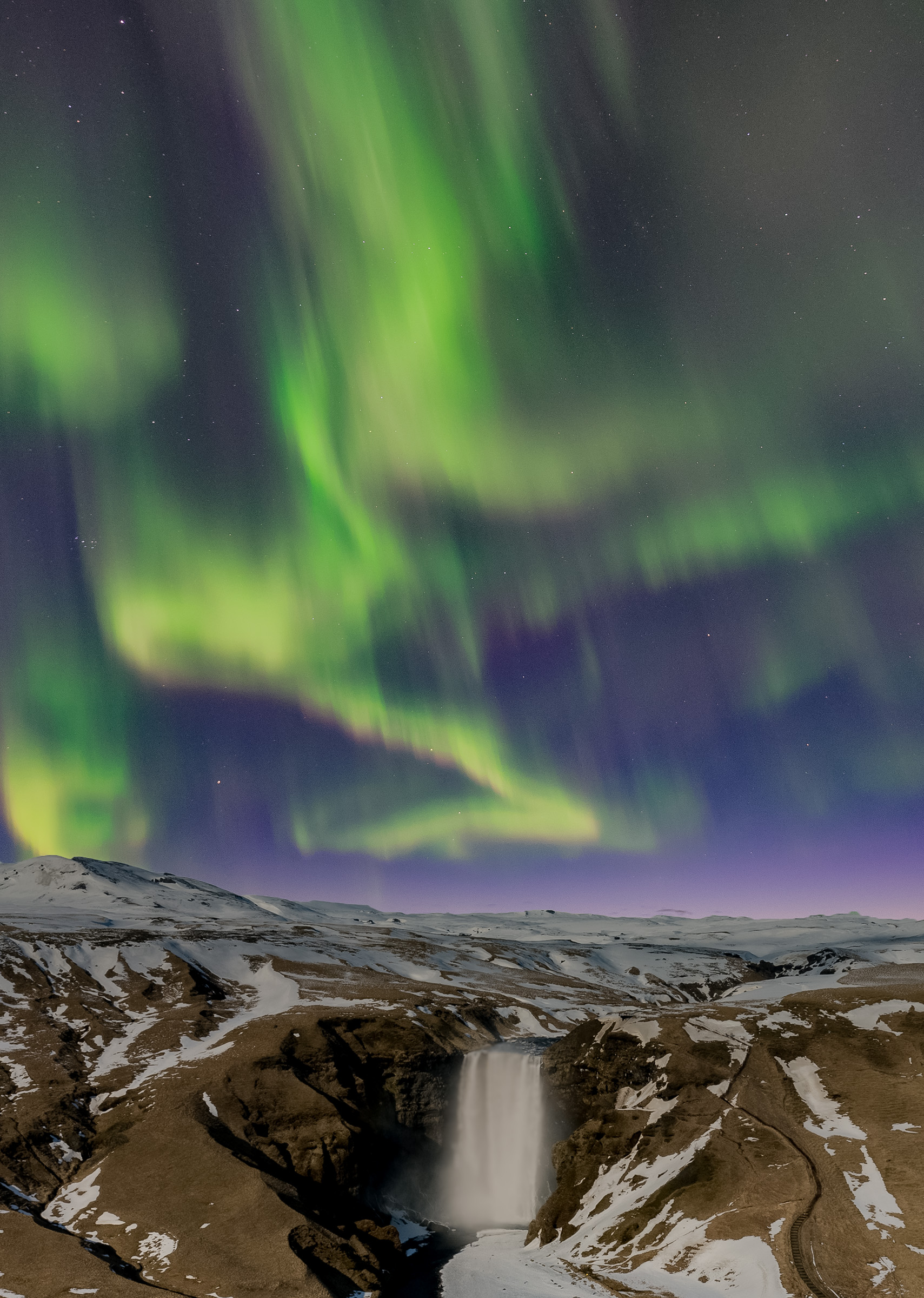 bright pink and green northern lights reflect in a pond, a church can be seen in backdrop in the snowy landscape