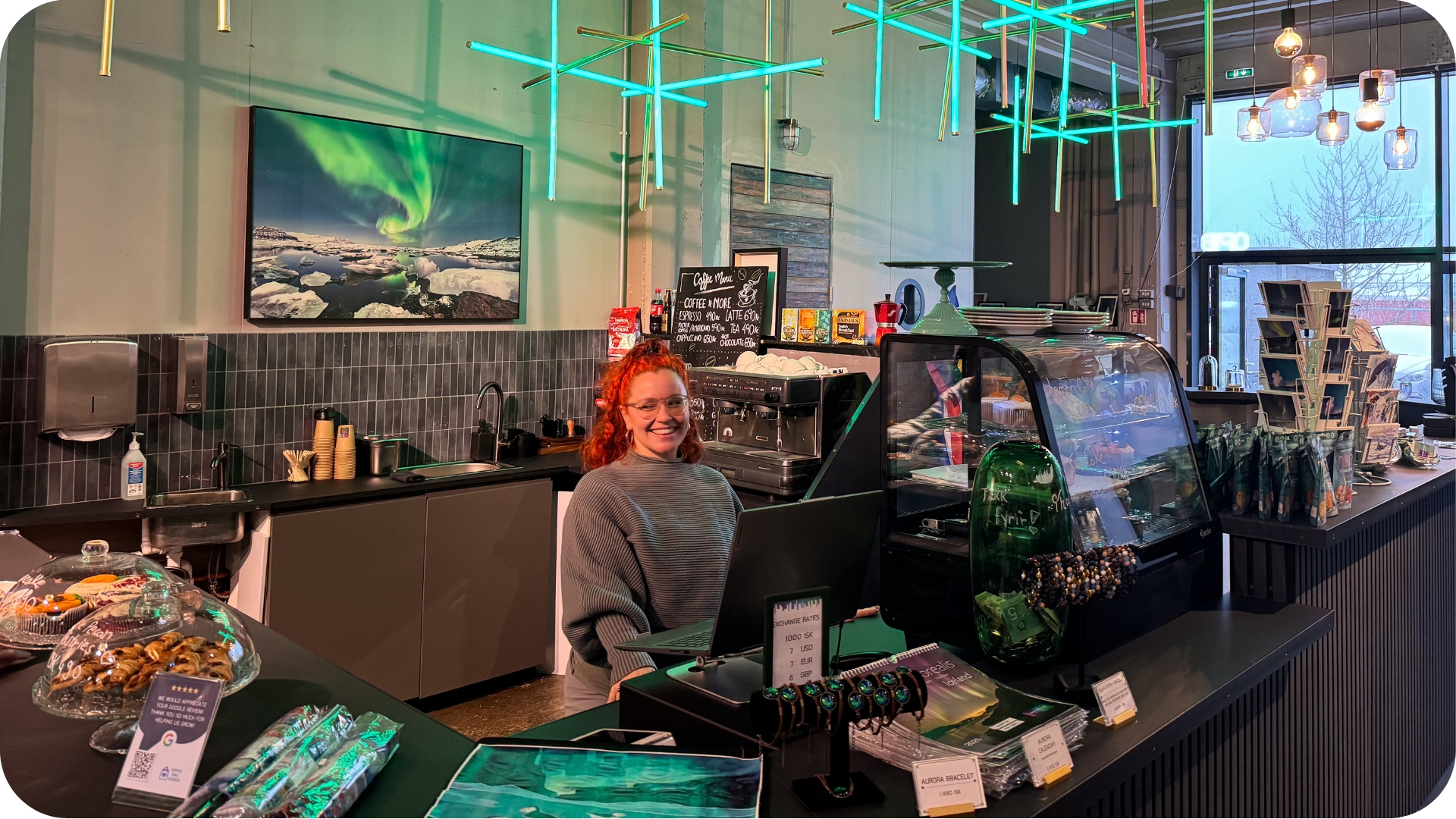 A smiling staff member behind the counter of the Aurora Reykjavík gift shop and café, surrounded by Icelandic souvenirs, aurora bracelets and handmade pastries, with northern lights-themed neon ceiling lights and a large aurora borealis photograph on the wall